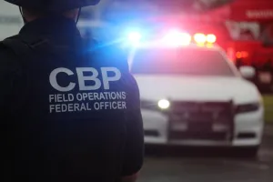 Rear view of CBP officer in uniform flanks vehicle with flashing lights.